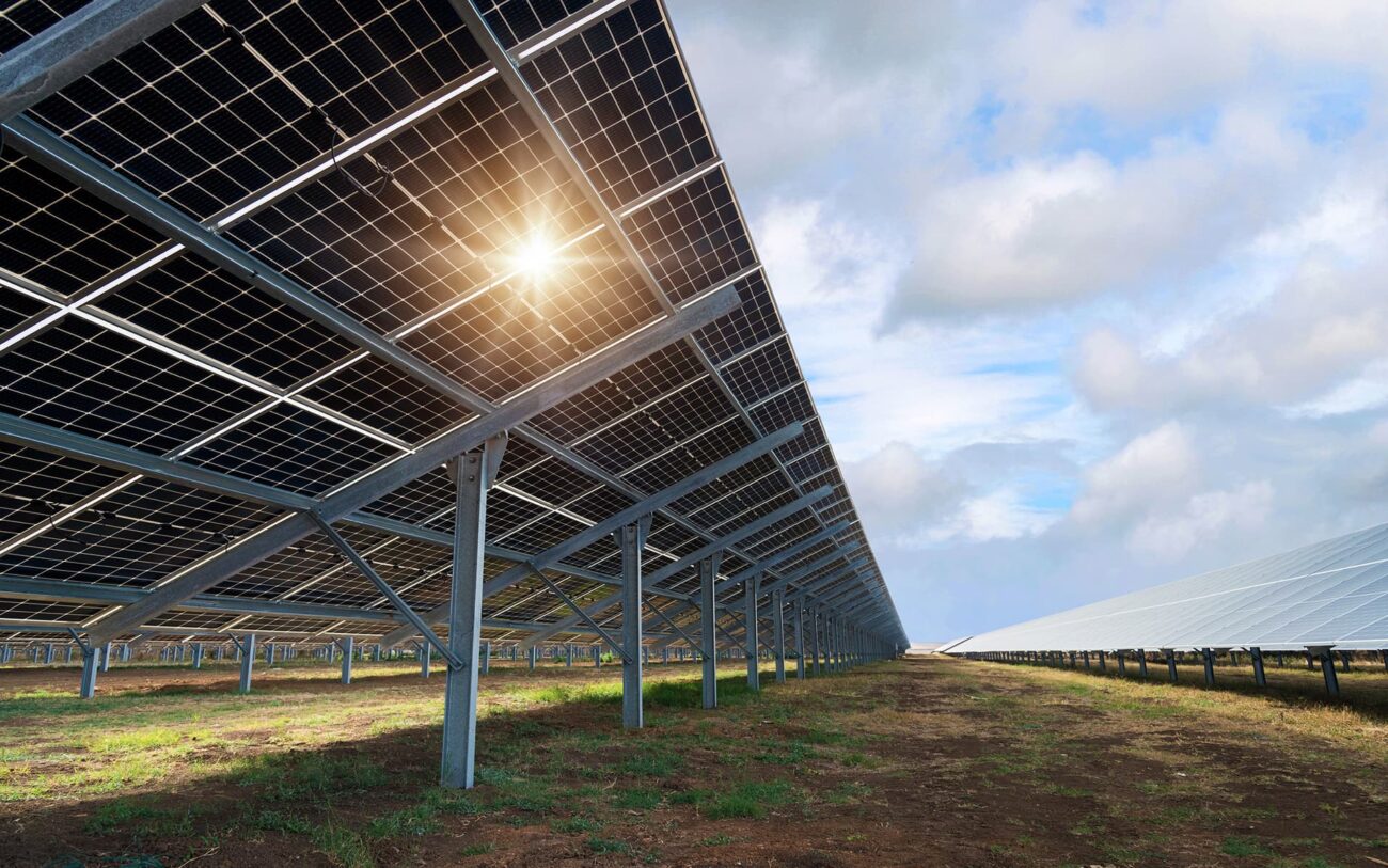 Aurinkopuisto, jossa on paljon maahan asennettuja aurinkopaneeleja. Paneelien välistä pilkistää aurinko ja sen säteet. Taustalla näkyy sinistä taivasta ja valkoisia pilviä.