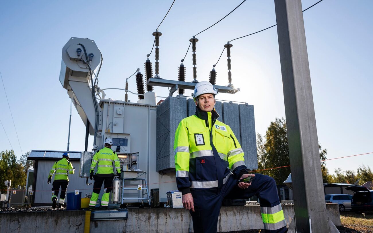 Oulun Seudun Sähkön työntekijä turvaliivissä ja kypärässä seisoo sähköinfrastruktuurin edessä.