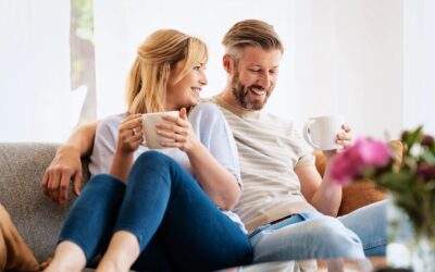 Kaksi henkilöä istuu rennosti lähekkäin sohvalla ja hymyilee toisilleen. Molemmilla on käsissään valkoiset kahvikupit. Etualalla näkyy sumeasti pinkkejä kukkia.
