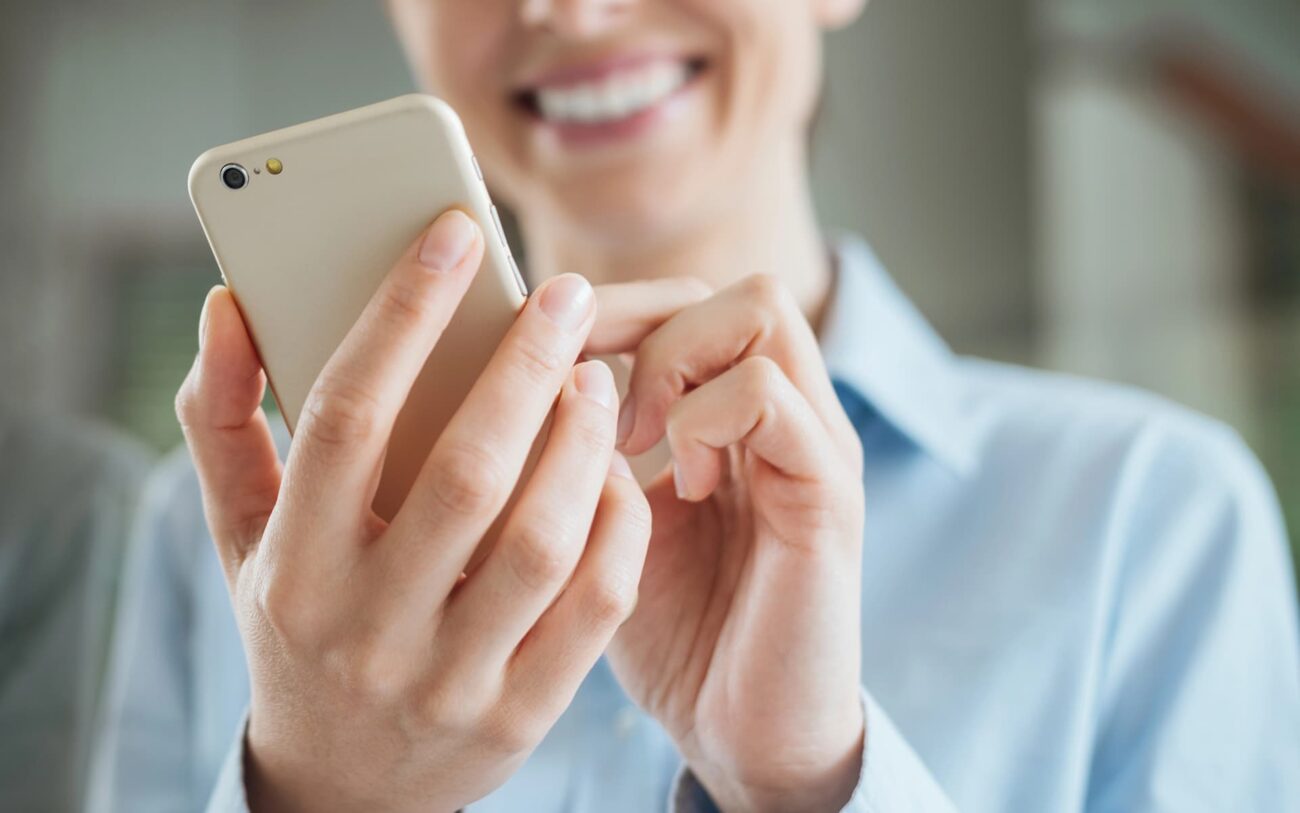 Henkilö pitää kädessään kännykkää ja käyttää sitä hymyillen. Kuvan tarkennus on henkilön käsissä ja kännykässä, henkilön kasvot näkyvät osittain sumeana taka-alalla.