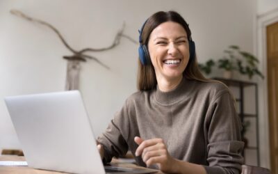 Henkilö käyttää kannettavaa tietokonetta kuulokkeet korvillaan ja hymyilee iloisesti sivulle katsoen.