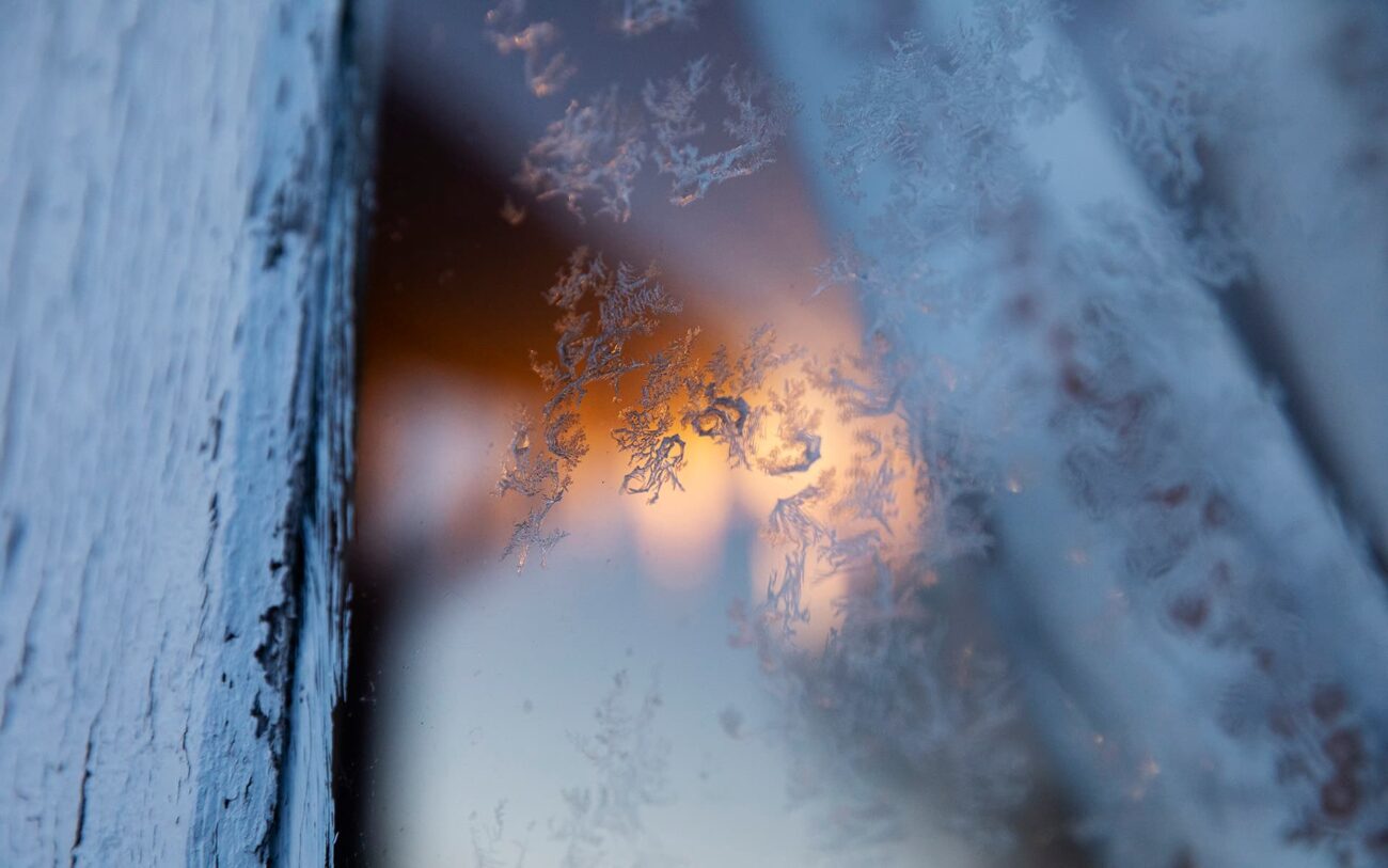 Kiteytyneitä lumihiutaleita talon ikkunassa. Talon sisältä loistaa lämmin valo.