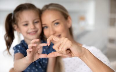 Perheen äiti ja nuori lapsi tekevät sormillaan sydämen ja näyttävät sitä kameralle. Tarkennus on henkilöiden käsissä ja niiden takana näkyy sumeasti henkilöiden hymyilevät kasvot.