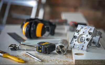 Lähikuva pöydällä olevista työkaluista, kuten mittanauhasta, suojakuulokkeista, maalauspensselistä ja kynästä. Pöytä ja tavarat ovat kevyen puupölyn peitossa.