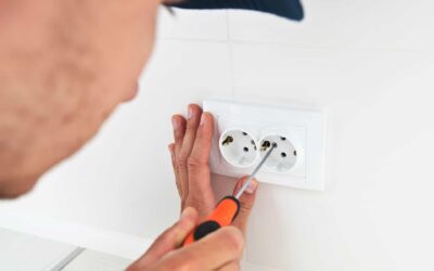 Sähköasentaja työssään ruuvaamassa pistorasian ruuveja auki. Asentajalla on päässään lippalakki ja kädessään ruuvimeisseli.