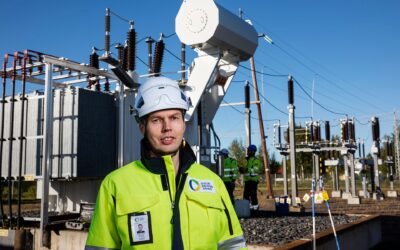 Oulun Seudun Sähkön työntekijä turvaliivissä ja kypärässä seisoo sähköinfrastruktuurin edessä.