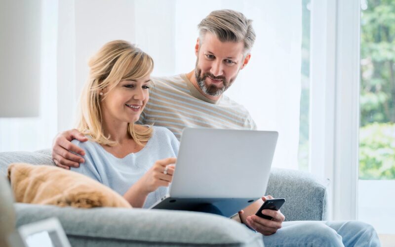 Kaksi henkilöä istuu lähekkäin sohvalla ja katsoo yhdessä kannettavaa tietokonetta, joka toisella henkilöllä on sylissään. Toinen henkilö osoittaa tietokoneen ruutua ja toisella on kädessään kännykkä.