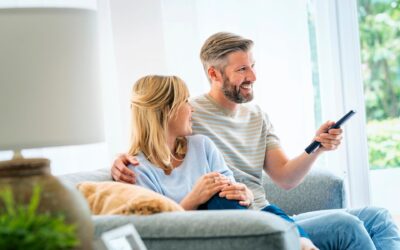 Kaksi henkilöä istuu lähekkäin sohvalla ja keskustelee keskenään. Toinen henkilöistä pitää kädessään kaukosäädintä ja kääntänyt kasvonsa oletettavasti television suuntaan.