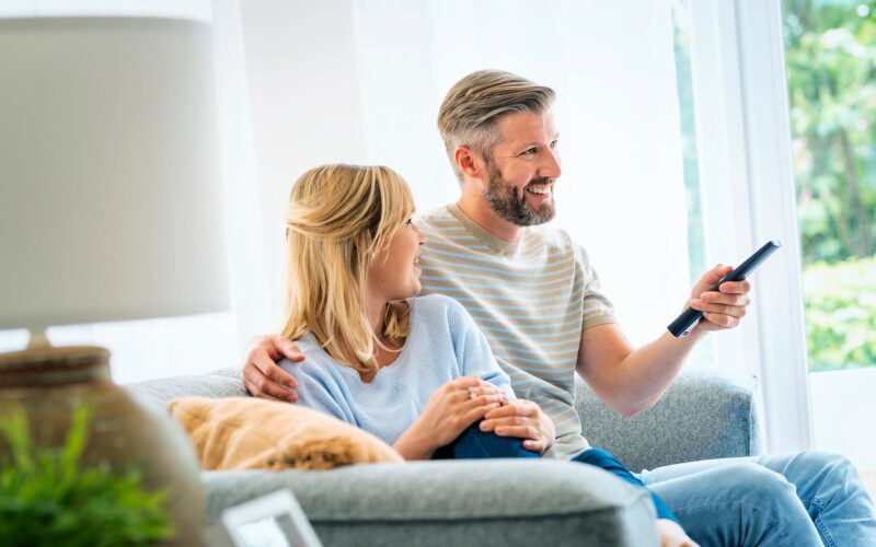 Kaksi henkilöä istuu lähekkäin sohvalla ja keskustelee keskenään. Toinen henkilöistä pitää kädessään kaukosäädintä ja kääntänyt kasvonsa oletettavasti television suuntaan.