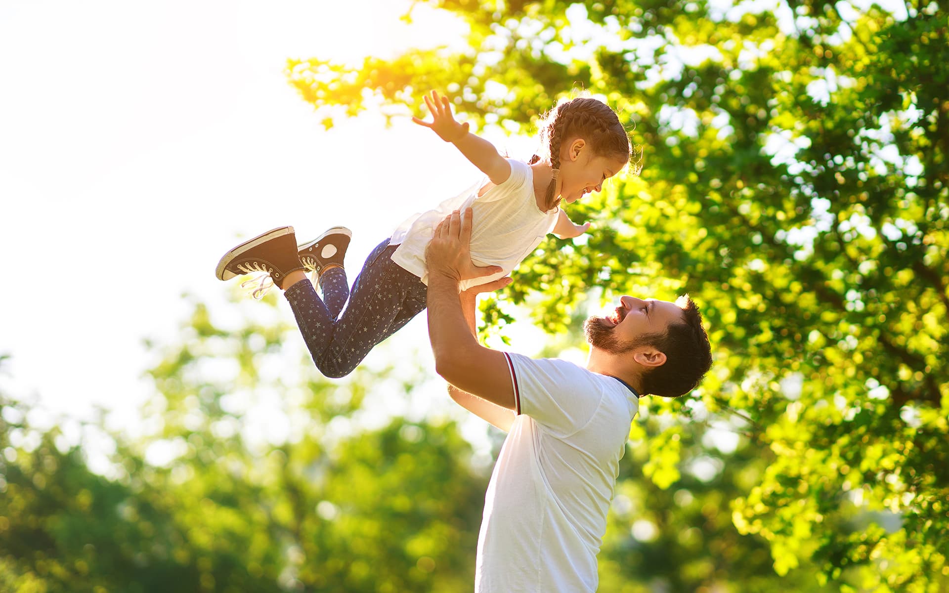 Perheen isä leikkii lapsen kanssa ja heittää häntä ilmaan. Lapsi levittää ilmassa kätensä ja katsoo hymyillen isäänsä. Henkilöt ovat ulkona ja taustalla näkyy vihreää puistoa sekä auringonpaistetta.