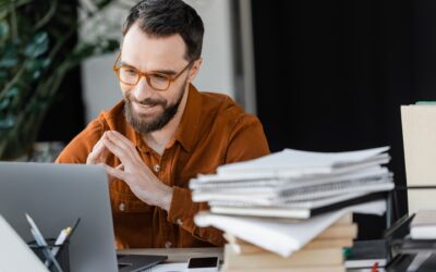 Henkilö katsoo hymyillen pöydällä olevaa, kannettavan tietokoneen näyttöä. Pöytä on täynnä papereita, vihkoja ja muuta tavaraa, kuten kyniä.