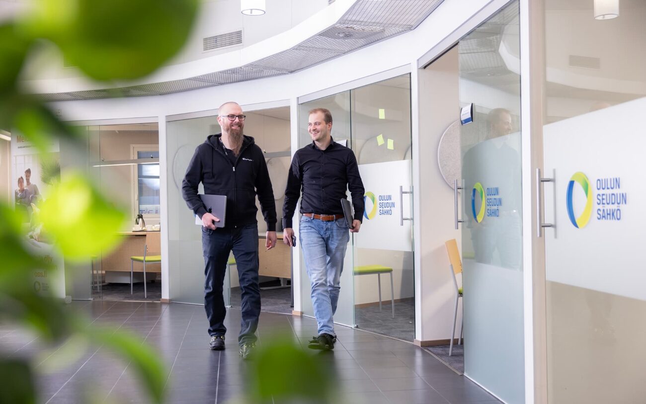 Kaksi työntekijää kävelee hymyillen Oulun Seudun Sähkön toimiston käytävällä. Molemmilla on mukanaan kannettava tietokone.