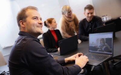 Oulun Seudun Sähkön neljä työntekijää kokoushuoneessa, työskentelemässä tietokoneilla. Etualalla mies katsoo oikealle hymyillen.