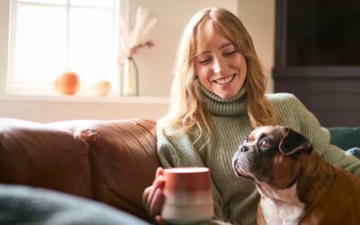 Henkilö istuu rennosti sohvalla, hymyilee ja pitää kädessään kahvikuppia. Henkilöllä on sylissään pieni koira, joka katselee sivulle. Taustalla näkyy sumeasti ikkuna ja ikkunalaudalle nostettuja koriste-esineitä.